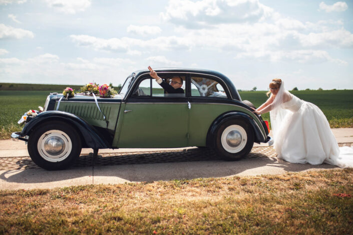 Die Braut im Hochzeitskleid versucht lachend, einen Oldtimer anzuschieben – humorvoller Moment, festgehalten von Christian Roder