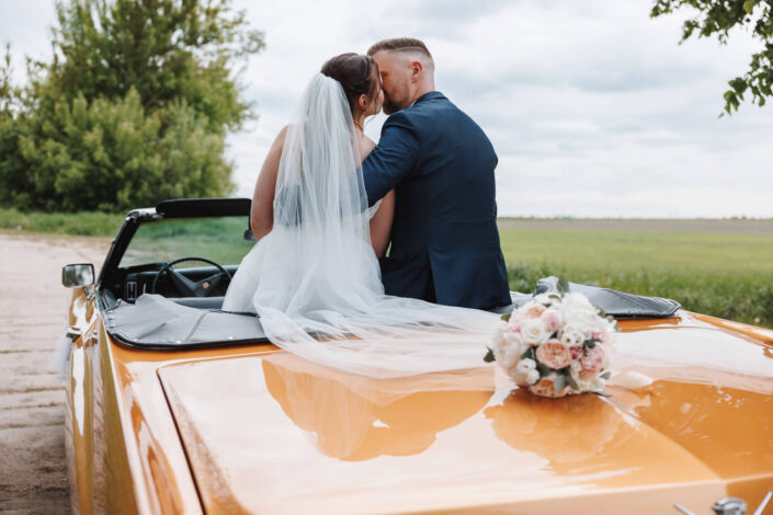Braut und Bräutigam küssen sich im orangenen Oldtimer, fotografiert von Christian Roder